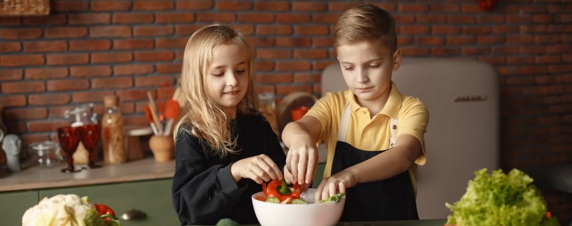 Cheerful girl helping little boy in apron preparing salad while standing in kitchen near table with mix of fresh vegetables behind brick wall